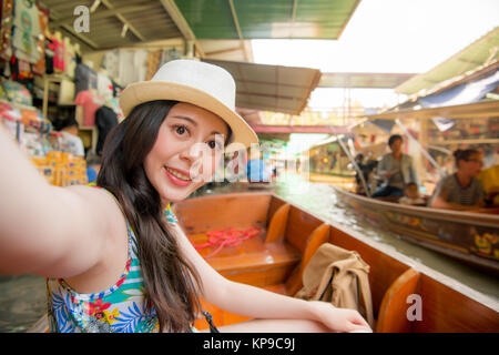 elegant attractive girl on river boat taking picture selfie and viewing floating market scene enjoying summer vacation during Thailand travel. Stock Photo
