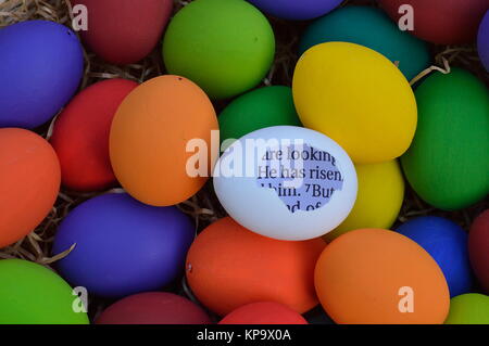 bright white egg with broken shell and bible text in it with the words: he has risen in the midst of many colorful easter eggs Stock Photo