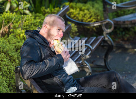 Man sitting outside eating a sub sandwich (large long bread roll or baguette) for lunch, in the UK. Stock Photo