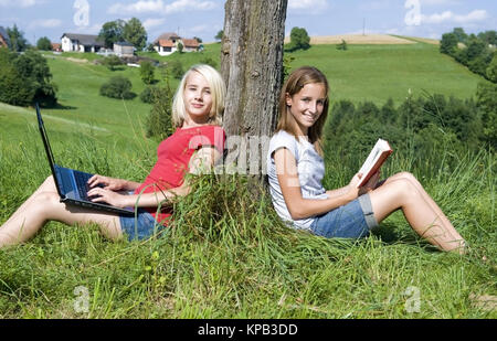 Model release, Zwei jugendliche Maedchen sitzen an Baumstamm gelehnt mit Laptop und Buch in der Wiese - two teenage girls ouside using laptop and read Stock Photo