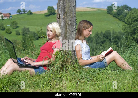 Model release, Zwei jugendliche Maedchen sitzen an Baumstamm gelehnt mit Laptop und Buch in der Wiese - two teenage girls ouside using laptop and read
