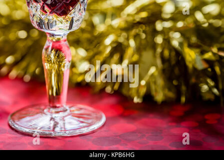 Glass of red wine detail, gold and red reflections on crystal. Christmas table closeup, gold tinsel decoration in background, side view. Stock Photo