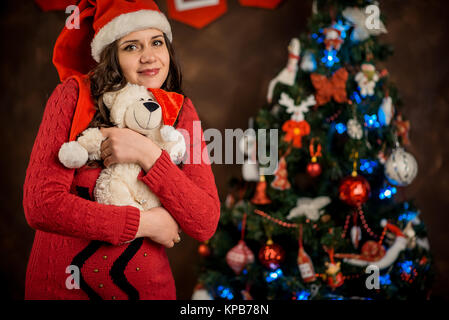 Horizontal portrait of the charming pregnant woman in Santa Claus hat hugging the lovely teddy bear at the background of the Christmas tree. Stock Photo