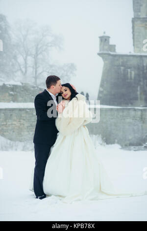 Romantic outdoor portrait of the happy newlywed couple. Groom is tenderly kissing his attractive brunette bride in forehead on the snowy meadow. Stock Photo