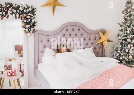 Portrait of cute funny little girl on Christmas morning in white home interior. Child pretending to be sleeping than rises cheerfully in bed. Horizont