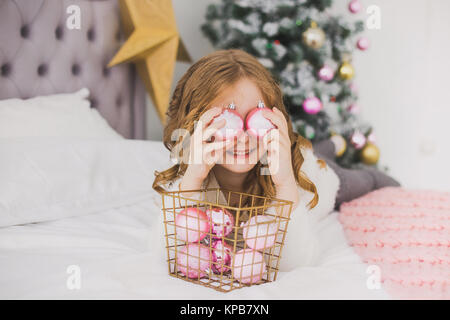Portrait of cute happy little girl on Christmas morning in home interior. Child paying with holiday decorations, making eye of pink balls while laying Stock Photo
