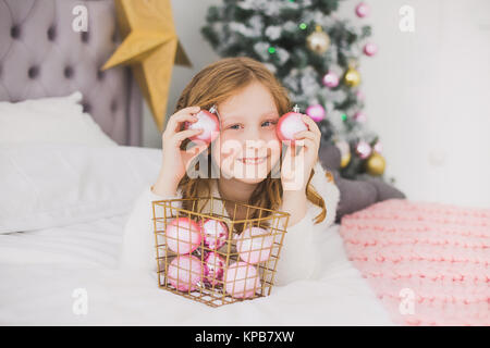 Portrait of cute happy little girl on Christmas morning in home interior. Child paying with holiday decorations, making eye of pink balls while laying Stock Photo