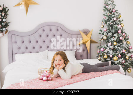 Portrait of cute happy little girl on Christmas morning in home interior. Child paying with holiday decoration while laying on bed. Horizontal color p Stock Photo