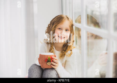 Portrait of cute happy little girl sitting on windowsill, drinking hot tea and looking outside from window. Child holding white mug in winter knitted  Stock Photo