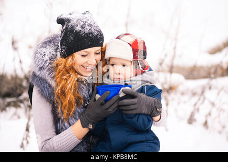 Baby sitting with young beautiful mother outside at snowy trees winter background and drinking hot tee. Happy family enjoying beautiful winter days on Stock Photo