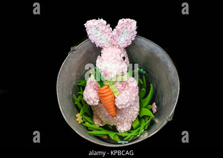 Pink Easter bunny made out of flower petals with yard carrot in a bucket with flowers isolated on black background Stock Photo