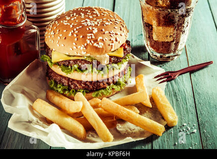 American double cheeseburger on a sesame bun served with crisp golden French fries, a soda or soft drink and ketchup as a fast food takeaway outdoors on a rustic table Stock Photo