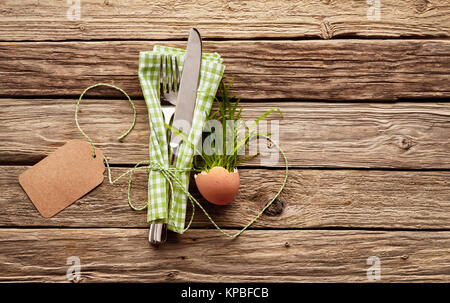 High Angle View of Country happy easter Themed Place Setting - Silver Knife and Fork Tied with String in Green and White Gingham Checkered Napkin Beside Plant in Eggshell Favor on Rustic Wood Table with Blank Tag Stock Photo