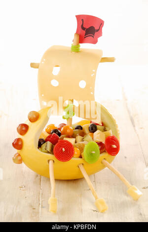 Creative food viking ship for a kids party made with fresh fruit salad, vegetables and cheese with a sail, flag, oars and shields down the hull displayed on a wooden table Stock Photo