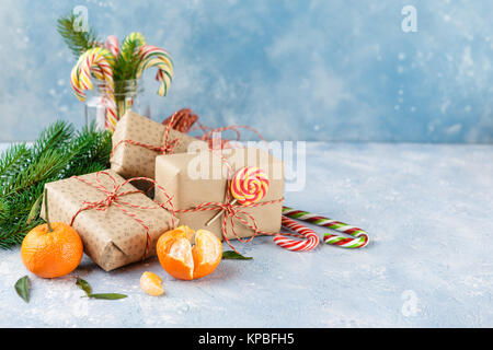 Gift boxes in kraft paper, fresh tangerines with leaves, candy canes and a branch of Christmas trees on a blue background Stock Photo