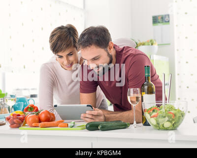 Young smiling couple cooking together at home, they are preparing vegetables and checking recipes online using a digital touch screen tablet Stock Photo