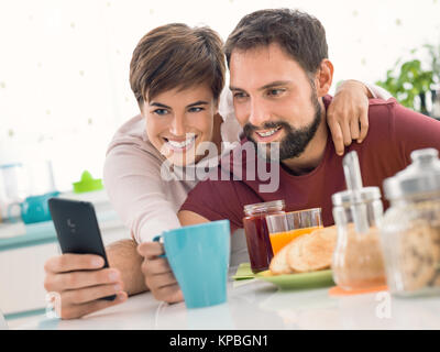 Young cheerful couple having breakfast at home together and taking selfies using a smartphone Stock Photo