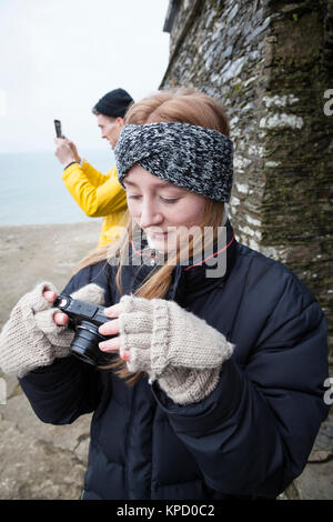 Girl dressed for Winter with bandana and gloves, checks her digital camera whilst behind a man takes a photo with his iPad, England. Stock Photo