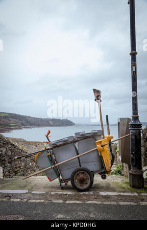 Street cleaner's trolley cart complete with brushes and shovel is parked by the side of the road with the sea in the background, Cawsand Stock Photo