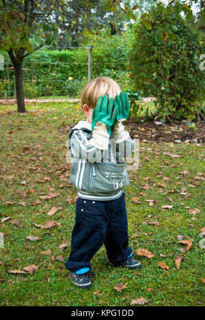 three year old boy hides behind adult gardening gloves in a game of hide and seek in a garden Stock Photo