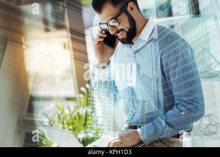 Bespectacled handsome man having phone conversation and using the laptop. Stock Photo