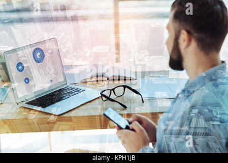 Young bearded man using cellphone and looking at the laptop. Stock Photo