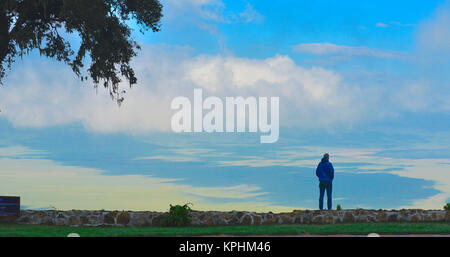 Man in blue standing on edge of Ngorongoro crater looking down into valley of green grass and white clouds. Landscape picture. Stock Photo