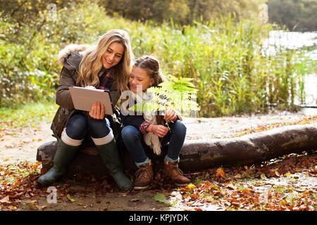 Mother using a digital tablet with her daughter sitting on a log. Stock Photo
