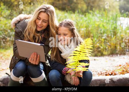 Mother using a digital tablet with her daughter sitting on a log. Stock Photo