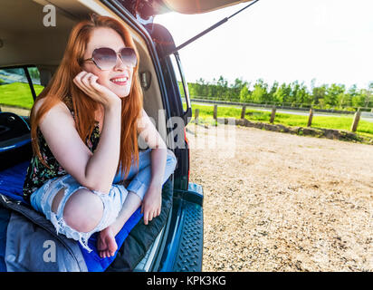 A young woman on a road trip lays in the back of a vehicle with her cell phone on a sleeping bag looking out from the  open door Stock Photo