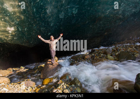 A man poses on a boulder with arms outstretched in front of a cave beneath the ice of Root Glacier in Wrangell-St. Elias National Park Stock Photo