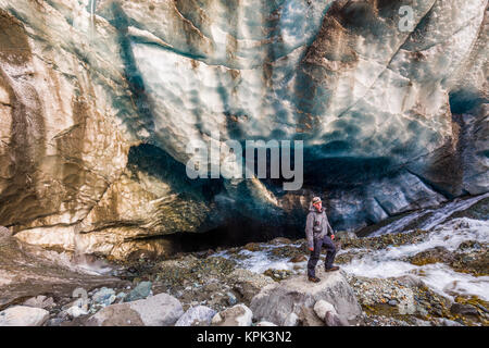 A man surrounded by rushing water poses in front of a cave beneath the ice of Root Glacier in Wrangell-St. Elias National Park Stock Photo