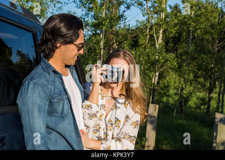 Young couple with camera standing outside their vehicle, the young woman taking a picture in the distance; Edmonton, Alberta, Canada Stock Photo