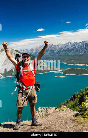 Female hiker on ridge top with arms in the air, and a colourful alpine lake and mountain range in the distance with blue sky and clouds Stock Photo