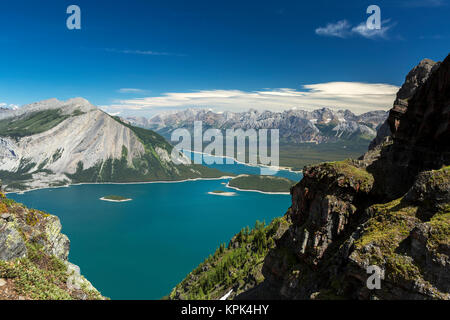 View from the top of mountain ridge looking down on colourful alpine lake and mountain range in the distance with blue sky and clouds Stock Photo