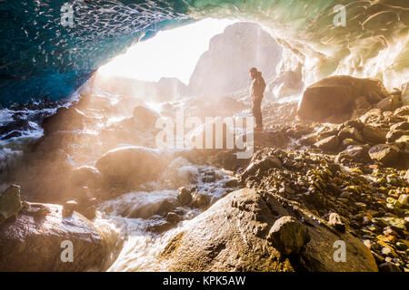 A man stands at the entrance of an ice cave bathed in warm sunlight and mist, Wrangell-St. Elias National Park; Alaska, United States of America Stock Photo