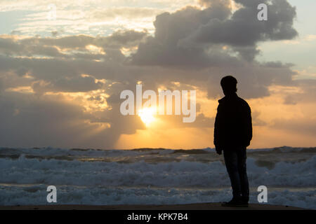 Rear view of a young silhouetted man standing on the beach at sunset Stock Photo