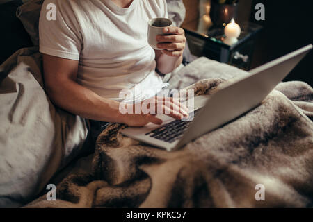Cropped shot of man on bed using laptop and having coffee.  Man working on laptop and drinking coffee on bed at home. Stock Photo