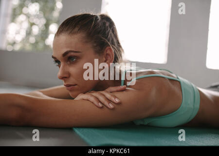 Close up of fit young woman taking rest after workout in health club. Female lying on exercise mat in gym and looking away. Stock Photo