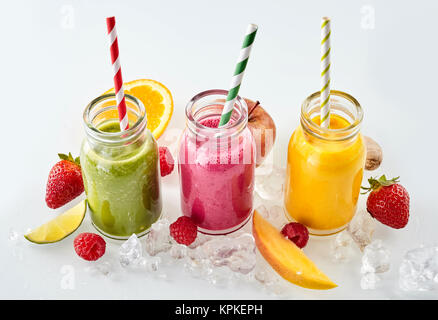 Row of apple, mango and strawberry smoothie beverages and matching straws surrounded by ice cubes and slices of fruit over gray background Stock Photo