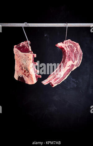 Two pieces of recently butchered raw uncooked cote de boeuf and tomahawek steak hanging from hooks on meat rack with copy space over black background Stock Photo