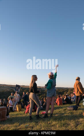 Large group of young, colorfully dressed music festival goers watching a magical golden sunset on the horizon with wind turbines in background. Stock Photo