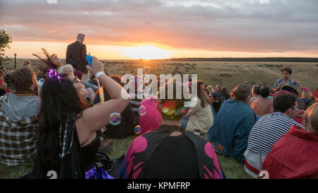 Women sitting in crowd of music festival goers, watching golden sunset while blowing soap bubbles. Stock Photo
