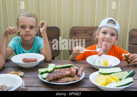 Two little girls at a wooden table in nature eating grilled sausages Stock Photo