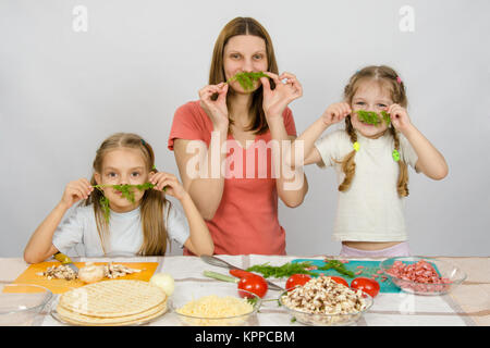 Young housewife with two daughters having fun holding sprig of parsley as a mustache at the kitchen table when sharing cooking Stock Photo