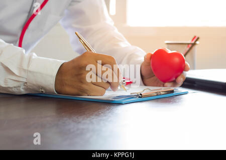 Doctor writing prescription on paper and holding red heart on working table Stock Photo