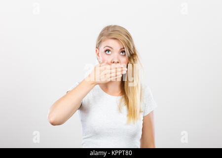 Saying bad things, shocking news concept. Ashamed young blonde woman having hand on her mouth. Studio shot on grey background. Stock Photo