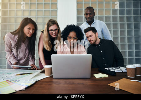 Smiling group of diverse work colleagues talking and working together on a laptop at a table in a modern office Stock Photo