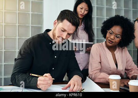 Young male architect working on a blueprint at a desk in a modern office with colleagues in the background Stock Photo