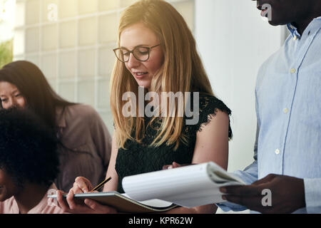 Two business colleagues writing down notes on clipboards while working in a modern office with colleagues in the background Stock Photo